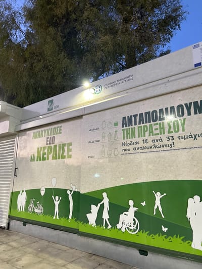 This image captures a recycling vending machine located outdoors in Koridallos, Greece, likely during dusk or early night, indicated by the dark blue sky and the illumination provided by a light fixture on the machine.
The machine, primarily off-white with green accents, is covered with informational text and graphics. On its left side, logos for the EU and the Hellenic Republic are visible, along with text identifying the initiative under "ΕΛΛΗΝΙΚΗ ΔΗΜΟΚΡΑΤΙΑ," "ΠΕΡΙΦΕΡΕΙΑ ΑΤΤΙΚΗΣ" (Region of Attica), and "ΕΔΣΝΑ" (a waste management authority). Bold green text in the lower section encourages users to "ΑΝΑΚΥΚΛΩΣΕ ΕΔΩ ΚΑΙ ΚΕΡΔΙΣΕ" (Recycle Here and Win).
The central and right panels feature a vibrant green landscape with white silhouettes of diverse people, including children playing, adults, a person in a wheelchair, and a family group, symbolizing inclusivity and community engagement in environmental efforts. Above these graphics, the machine states "ΑΝΤΑΠΟΔΙΔΟΥΜΕ ΤΗΝ ΠΡΑΞΗ ΣΟΥ" (We Reward Your Action). Further details explain the reward system: "Κέρδισε 1€ ανά 33 τεμάχια που ανακυκλώνεις!" (Win 1€ for every 33 items you recycle!), with redemption possible through free purchases at collaborating stores. Icons and labels for accepted recyclable materials are displayed: "ΜΕΤΑΛΛΟ" (Metal), "ΠΛΑΣΤΙΚΟ" (Plastic), and "ΓΥΑΛΙ" (Glass).
The overall scene depicts a public service encouraging recycling through a reward mechanism, set against a backdrop of trees and a dark sky. A closed metal shutter is visible at the bottom right of the machine.