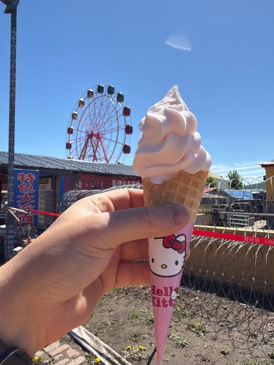 This outdoor photograph, set in Jiejinkou, China, captures a person's right hand holding a light pink soft-serve ice cream cone. The ice cream is swirled high above a crisp waffle cone, which is encased in a decorative paper sleeve featuring the iconic "Hello Kitty" character and logo. "Hello Kitty" is clearly visible in English text on the wrapper.

In the background, against a clear, bright blue sky, a large red Ferris wheel with colorful gondolas stands prominently. To the right, a security fence constructed with barbed wire runs horizontally, interwoven with a distinct red ribbon or tape. Below the Ferris wheel, several buildings and signs bearing blurry Chinese characters are discernible. The foreground shows unpaved ground with scattered dirt and small plants, suggesting a casual, perhaps fairground-like setting. The overall scene indicates a sunny daytime environment, likely a leisure or amusement area.