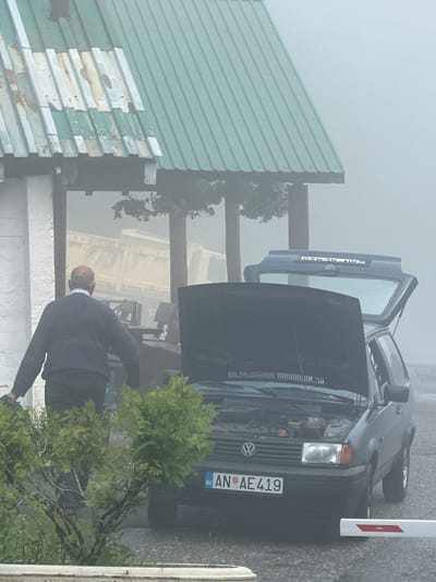 In Trešnjevik, Montenegro, amidst a dense fog, an older man wearing a navy blue sweater and dark pants stands near a dark-colored Volkswagen hatchback. The car's hood is open, revealing its engine. The rear hatch of the car is also open. A license plate on the front of the car reads "AN AE 419". The scene takes place outdoors, with a building featuring a green corrugated metal roof to the left. Vegetation, including bushes and trees, is visible in the foreground. The overall atmosphere is hazy and obscured by fog.