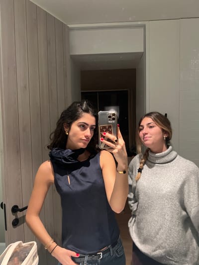 Two women pose in a mirror selfie. One wears a grey turtleneck sweater and has her hair in a braid. The other holds the phone, wearing a navy blue top, jeans, and a belt with a silver buckle. She has dark hair and wears gold jewelry. The background includes a white wall, a doorway leading to a dark room, and a light-colored wooden surface. The phone has a clear case with a decorative card inside.