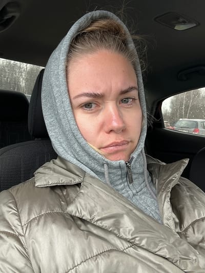 A woman with blonde hair is in a car in Ural'skoe, Russia, wearing a gray hoodie and a beige puffer jacket. Snowflakes are visible on the car window. The car seat is black.