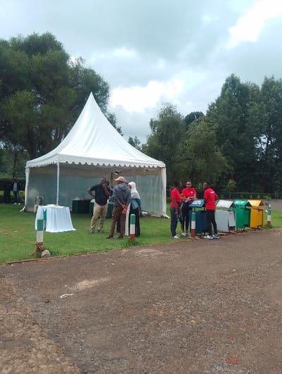 This outdoor image captures an event or gathering in a green, park-like setting under an overcast sky, likely in Gitaru, Kenya. A prominent white, pyramid-shaped tent stands in the center-left, with a table draped in a white tablecloth visible underneath.
Several individuals are present. On the left, near the tent, a few men are conversing; one wears a black shirt, and another wears a light-colored hat. A person in a white hijab is also standing near this group. To the right, a group of four people, possibly young adults, are wearing matching red shirts. They are gathered near a series of colorful recycling bins – green, yellow, and blue. One of the blue bins clearly displays the text "PAPER WASTE."
The foreground features a rough, paved road or path. Green grass covers the ground where the tent and people are situated, bordered by the paved area. Green and white striped bollards are visible, delineating sections of the ground. The background is dominated by lush green trees, suggesting a natural or well-maintained environment. The cloudy sky indicates daytime, likely a mild day without direct sunlight. The presence of recycling bins and the coordinated red shirts of the group on the right suggest an environmental awareness or community cleanup event. The overall atmosphere appears calm and organized.