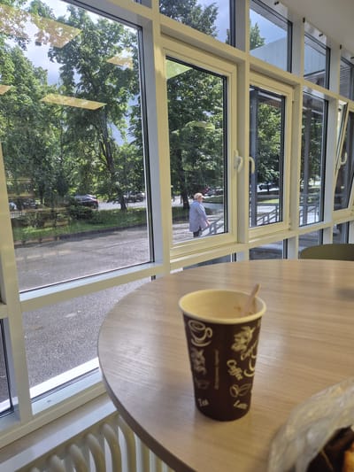 A paper coffee cup with a wooden stirrer rests on a light-colored wooden table. The cup is brown with white text and graphics, including the words "Coffee" and "Latte," and designs resembling coffee beans and leaves. In the background, a large window with multiple panes reveals an outdoor scene. Through the glass, trees with lush green foliage are visible, suggesting a daytime setting, likely during warmer months. The reflection in the window shows a person wearing a light-colored shirt and a hat, walking outside. The floor outside appears to be paved with asphalt. The overall setting suggests a cafe or an indoor seating area with a view of the outdoors. The image is oriented vertically.