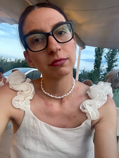 The image shows a close-up, self-portrait of a woman with dark hair and glasses. She is wearing a white ruffled dress and a pearl necklace. Her lips are painted with a dark lipstick. There are red marks on her chest, possibly from a rash or a hickey. The background suggests an outdoor setting with trees and a cloudy sky, indicating it's daytime. The orientation of the photo suggests the woman is holding the camera above her head. The setting appears to be casual, possibly a social gathering or an outdoor restaurant. The image is a personal photograph, likely taken by the subject herself, and the focus is on her face and upper body. No visible text is present. The overall mood of the image is unclear due to the close-up and the subject's expression, though the red marks on her neck could suggest a sense of discomfort or arousal.