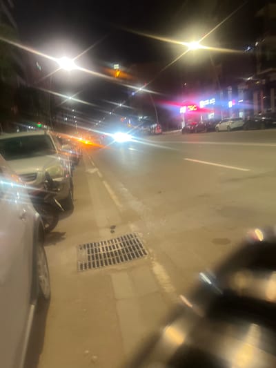 A nighttime urban street scene, possibly captured from a moving or parked vehicle, depicts a street in Beni Mellal, Morocco. The image, if rotated 90 degrees counter-clockwise, shows a wet or reflective asphalt road surface on the left, with faint white lane markings and a dark square drainage grate on the curb. The right side of the frame features blurry elements of the vehicle from which the photo was taken, including part of a white hood or roof and a side mirror, along with reflections on a windshield.
Along the left side of the street, buildings or shops are brightly illuminated by numerous artificial light sources, including streetlights and various shop signs, which create prominent lens flares and starburst effects. Several vehicles, including a white car, are visible on the street and parked alongside. Illuminated signs with visible Arabic script, notably one in bright red and another in blue, confirm the Moroccan setting. The dark sky and reliance on artificial lighting clearly indicate it is nighttime. The damp appearance of the road suggests recent rain or wet conditions.