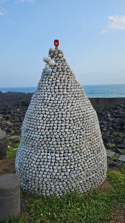 The image shows a large, conical structure constructed from numerous small, white shells, resembling a haystack or a beehive. The structure is situated outdoors, on a grassy area with dark volcanic rocks scattered around it. In the background, the ocean is visible, with a calm blue surface and a grey, low wall or barrier along the coastline. The sky above is a light blue with a few wispy clouds.
At the very top of the shell structure, there is a red and gold finial, possibly a light or marker. Several white buoys and a single light blue buoy are attached to the upper portion of the structure, draped with a green rope.
This structure is located on Jeju Island, specifically in the city of Seogwipo-si, South Korea. It is likely part of a coastal art installation or a cultural landmark. There are no people visible in the image, nor any actions or interactions taking place. The scene is static and focuses on the large shell sculpture and its natural, seaside environment.