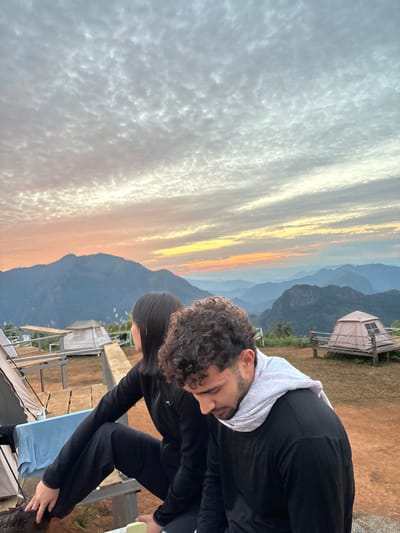 Two individuals are seated on a wooden platform at a mountainous campsite in the vicinity of Nong Ian, Laos, during either sunrise or sunset. The sky displays scattered altocumulus clouds, illuminated by orange and yellow light along the distant horizon, transitioning to grey and white overhead. A female, with dark, straight hair, wearing a black long-sleeved top and pants, sits with her back to the camera, looking towards the layered mountain ranges in the distance. To her right, a male with dark, curly hair, dressed in a black long-sleeved shirt and a light-colored scarf, sits with his head bowed downwards. In the background, at least three light-colored dome tents are visible on elevated wooden platforms, interspersed among sparse vegetation on the earthy terrain. Multiple receding mountain ridges, covered in dark green foliage, extend into the hazy distance under the twilight sky.