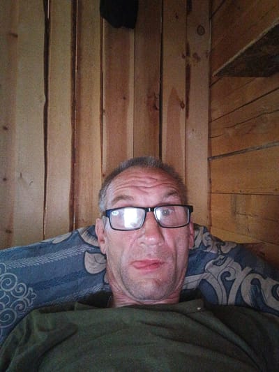 A middle-aged man with short graying hair and wrinkles on his forehead is lying down, resting his head on a patterned pillow. He is wearing black-rimmed glasses with a faint prescription sticker on the left lens and a dark green t-shirt. The background consists of wooden planks, suggesting an indoor environment, possibly a room with a rustic aesthetic. The lighting is warm and somewhat dim. The scene does not provide enough information to determine the specific location in Strelka, Russia.