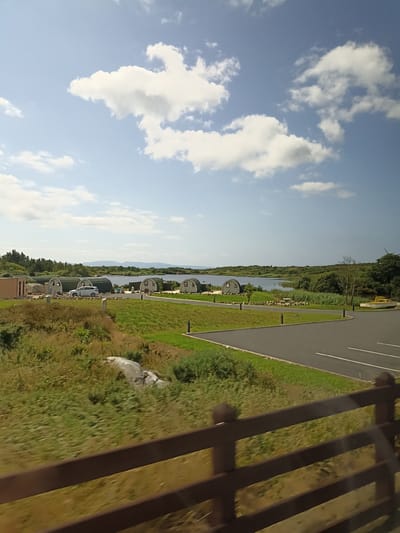 The image shows a scenic view of a resort or camping site in Burtonport, Ireland, on a bright, sunny day. In the foreground, a wooden fence runs horizontally across the frame, with blurred green grass and foliage behind it.

The middle ground features a neatly paved asphalt road with white parking lines, bordering a well-maintained grassy area. Several dome-shaped accommodations, likely pods or cabins, are visible. A white car is parked near one of these structures. In the background, a body of water, possibly a lake or bay, stretches towards the horizon, with lush green trees and vegetation lining its banks. Distant hills or mountains are faintly visible against the horizon. The sky is a clear blue with scattered white clouds. A yellow boat is docked on the shore near some of the accommodations.