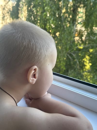 A young child, seen from the side, with light hair and fair skin, rests their chin on their hands while looking out a window. The background is a blur of green foliage and sunlight, suggesting an outdoor scene beyond the window. The child is wearing a thin dark cord necklace. The windowsill is white. The image was taken in the Yarominski Sel'ski Sovet, Belarus.