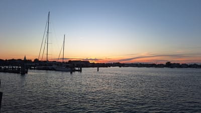 This is a photograph of a harbor on Nantucket, USA, at sunset. The sky transitions from a clear, pale blue at the top to a vibrant orange and pink hue near the horizon, with wispy clouds catching the light. Two sailboats, silhouetted against the sunset, are moored in the foreground, their tall masts dominating the left side of the image. A dock with pilings extends into the water from the left edge. In the middle ground, a line of buildings, some with gabled roofs, stretches along the shoreline. The water in the harbor is a dark, rippling surface, reflecting the muted colors of the sky. Other boats are visible anchored further out in the harbor, and the silhouette of a church spire can be seen among the buildings on the left. The overall scene evokes a peaceful, end-of-day atmosphere.