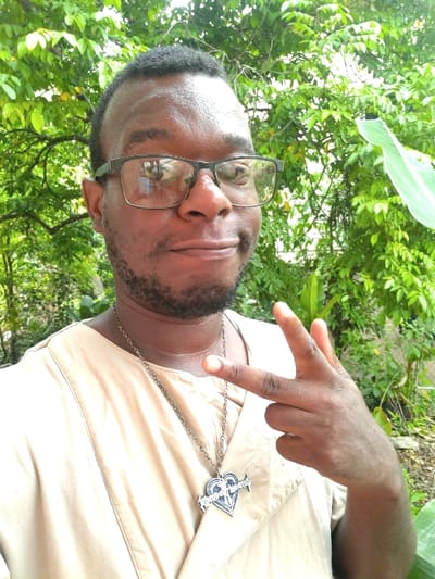 The image shows a man standing outside in La Romaine, Trinidad and Tobago, in front of a lush, tropical environment. The man is wearing a beige shirt and a silver necklace with a heart-shaped pendant. He has short dark hair and is wearing black-framed glasses. His left hand is extended outward with the index and middle fingers extended. He is smiling with a slightly tilted head. The background is a green, leafy landscape with large leaves and a hint of a wall in the distance. The man's expression is relaxed and his posture is casual.