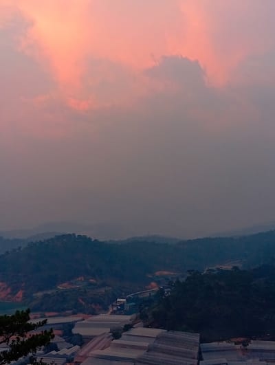 The image depicts a landscape in Unknown, Vietnam, featuring a hazy, pink-tinged sky above layers of dark, rolling hills. Greenhouses dominate the foreground, their roofs arranged in tiers. A road winds through the hills, leading to a cluster of buildings. The scene appears to be viewed from an elevated position.
