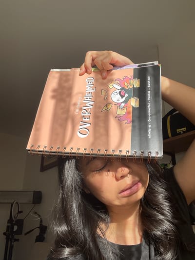 A person is holding a notebook over their face, obscuring it. The notebook has a pink cover with the words "I'm feeling OVERWHELMED" printed in large, white letters. A cartoon character is depicted on the notebook, looking distressed amidst scattered papers and flames. To the right of the notebook, a black and yellow Nikon camera bag is visible on a shelf. The person's dark hair falls around their face, and sunlight casts a shadow pattern on their skin. The background suggests an indoor environment with a microphone stand and other equipment visible. The scene does not depict any specific location in Başakşehir, Turkey.