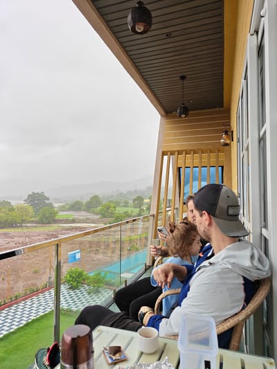The image depicts a family enjoying a view from a balcony in Ajivali, India. A man wearing a baseball cap and a blue and white jacket is seated in a wicker chair, looking towards the right. Next to him, a young girl with curly hair is also seated, holding a glass. Another man is visible behind them, holding a smartphone. The balcony overlooks a rural landscape with hills in the distance, under a cloudy sky. In the foreground, a glass-topped table holds a mug, a small plate with food, and a plastic container. The balcony is furnished with a railing and a tiled floor pattern.
