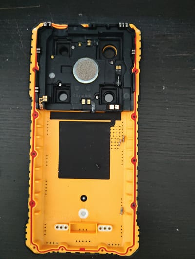The image shows the disassembled back casing of a rugged smartphone, lying on a dark, textured surface that resembles wood. The casing is primarily bright yellow with a red rubber seal along the edges. The left side of the casing, painted black, houses several components. These include a circular speaker with a textured gray mesh, and two smaller circular openings, likely for buttons or other hardware. There are also several small screws and connectors visible. Some text is printed on the black component: "ASUS_MAIN_FR DL_24" and "G05006".
The right side of the casing is predominantly yellow and appears to be the main housing area. A rectangular black adhesive pad is adhered to this section. There are several small holes arranged in rows, possibly for ventilation or mounting. More text is printed on this yellow surface: "GD1008 PC+10%GF". The overall scene suggests a repair or disassembly process, with the internal components of the phone exposed. The lighting is consistent, suggesting it was taken indoors under artificial light. There are no people or indications of weather. The location context of Mělník, Czechia, is not visually discernible from the image itself.