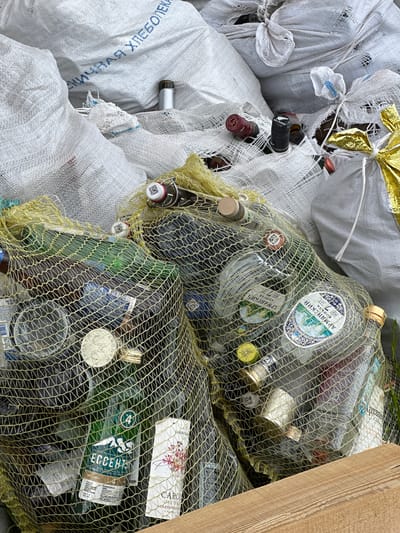 The image displays a close-up, high-angle view of a pile of collected glass bottles, predominantly stored in bags, situated in Kuda, Russia. The scene suggests a waste collection point for recyclables.
Numerous bottles of various sizes, shapes, and colors (clear, green, and brown) are visible. Many are contained within large, yellowish-green coarse mesh bags, which allow their contents to be seen. Other bottles are held in white, woven polypropylene bags, some of which appear torn or worn. A decorative golden, metallic-looking bow adorns one of the white bags. A wooden plank is visible along the right side of the frame. The bright, diffuse lighting indicates it is daytime. No people are present, depicting an unstaffed collection area.
Several labels and inscriptions on the bottles are discernible. Bottles for "АРМЯНСКИЙ КОНЬЯК" (Armenian Cognac) are visible, with indications of "3 ГОДА" (3 years) and "7 ЛЕТ" (7 years) on their labels. A green bottle is identified as "ЕССЕНТУКИ" (Essentuki) natural mineral water, labeled "ВОДА МИНЕРАЛЬНАЯ ПРИРОДНАЯ". Other labels include "MAMONT" on a circular, metallic tag, and fragments of a wine label such as "CAROL RESE CABERNET SANTA CA...". A black bottle cap reads "JOHN J. SAINSBURY LTD", and another states "NEW ZEALAND". On one of the white woven bags, the Cyrillic text "ХЛЕБОПЕКА" (Khlebopek - Baker/Bakery) is visible, indicating its prior use, likely for flour. Various barcodes and QR codes are also present on caps and labels.
