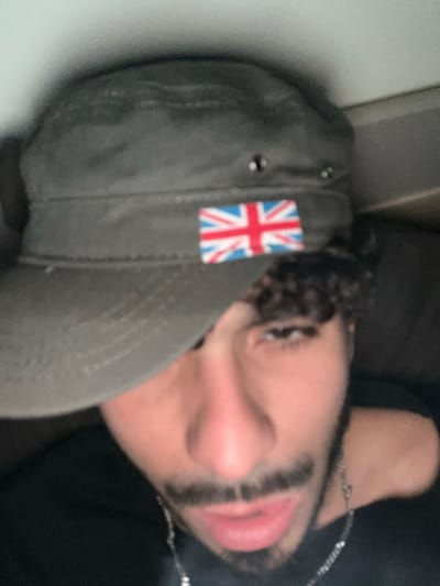 The image is a blurry, close-up portrait of an individual located in Marcoussis, France. The subject, appearing to be male, faces the camera with a direct gaze. They wear a dark olive-green military-style cap, featuring a Union Jack flag patch prominently displayed on the front left side. Two metallic grommets are visible on the cap's side. Dark, curly hair is visible beneath the cap on the subject's right side. The individual has a dark mustache and beard, and their lips are slightly parted. A silver-colored chain necklace is visible around their neck. The subject is wearing a dark-colored top, possibly black. The background is dark and out of focus, providing no discernible environmental details, suggesting an indoor setting with limited illumination. No specific actions or interactions are evident beyond posing for the photograph.