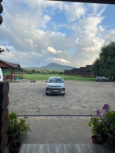 A modern, silver SUV is parked on a stone-paved courtyard in front of a rural establishment. The SUV's license plate, K575HT 126, is visible. To the left of the SUV, a small, black toy police car is placed on the ground. In the background, a covered wooden structure with tables and chairs is situated. Further back, a row of wooden houses with dark roofs stretches across a green field. In the distance, a mountain range with lush green slopes under a cloudy sky dominates the horizon. The scene appears to be set in the city of Dakhovskaya, Russia. Potted plants with purple and red flowers are visible in the foreground.