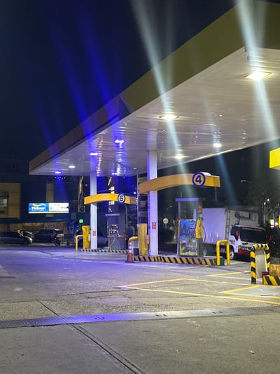 At a gas station in Perímetro Urbano Barranquilla, Colombia, at night, a wide shot captures the illuminated canopy extending over multiple gas pumps. The canopy's underside features white reflective panels and bright, square lights, while the edges are lined with yellow trim. Blue lights are prominently displayed, casting beams onto the dark sky and the canopy's structure. To the left, a "Pintuco" billboard is visible, illuminated with white and blue lights. Several cars are parked and in motion on the asphalt forecourt, which is marked with yellow lines. Gas pumps, some with orange safety cones nearby, are positioned under the canopy. One pump displays a blue circle with the number "6" inside, and another pump has a blue circle with the number "4". A white truck with red accents is parked to the right, and a person is seen standing near a pump. The surrounding area appears to be urban, with buildings and trees visible in the background.