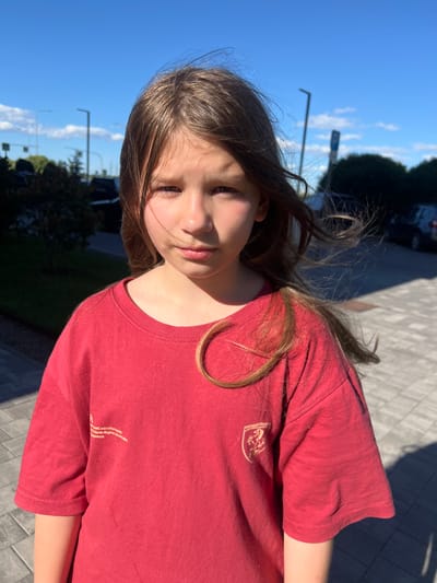 A young girl with long, light brown hair is looking towards the camera, her face slightly turned to the side. She is wearing a red t-shirt with a small emblem on the left chest and some faint text on the lower right side of the shirt. The t-shirt is short-sleeved. The girl appears to be outdoors, possibly in a park or on a paved pathway. The sun is shining brightly, casting shadows and highlighting her face and hair. The sky is a clear blue with some scattered white clouds. Trees and some streetlights are visible in the background, suggesting an urban or suburban setting. The overall mood is casual and relaxed, with the girl's expression conveying a sense of curiosity or slight shyness. The time of day is likely afternoon, given the strong sunlight and the angle of the shadows. The location context is Novoselye, Russia.