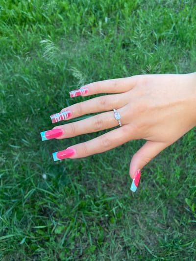 A person's left hand, with long, acrylic nails, is shown against a background of green grass. The nails are painted in a summer-themed design with pink and light blue colors. The ring finger wears a silver ring with a rectangular stone. The image was taken outdoors during daylight. The location is Askarovo, Russia.
