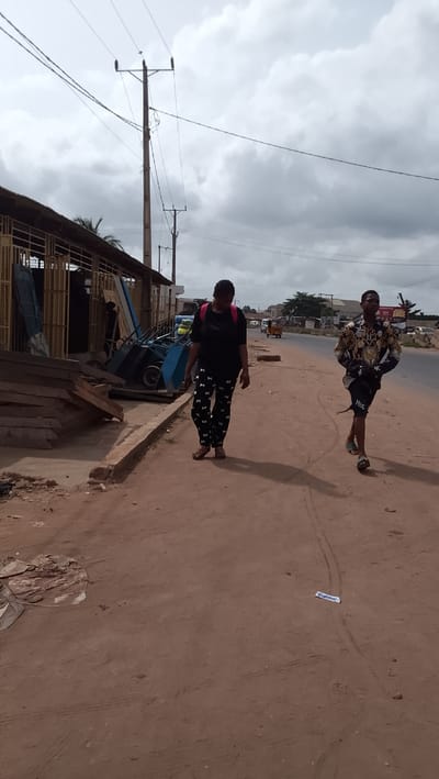 The scene is located in Ota, Nigeria, at the coordinates latitude: 6.683, longitude: 3.18. Two people are walking on a dirt road, which is on the edge of a paved road. The person on the left is a woman wearing a black shirt, black and white pants, and a backpack. She is walking towards the camera, and the other person is walking away from the camera on the right side of the image. The person on the right is a young man wearing a gold-colored shirt with a black and white pattern, black shorts, and sandals. The paved road behind them has a white lane divider line and appears to be a busy street. In the background, there is a variety of buildings and vegetation. On the left side of the image, there is a small building with a blue roof, wooden boards, and two large wheelbarrows. The building has a wire fence and appears to be a construction site or workshop. There is a power pole in the middle of the image with several electrical wires running across the sky. There is a tree on the left and a building on the right side of the image. The image is shot during the day, and the sky is overcast with white clouds.
