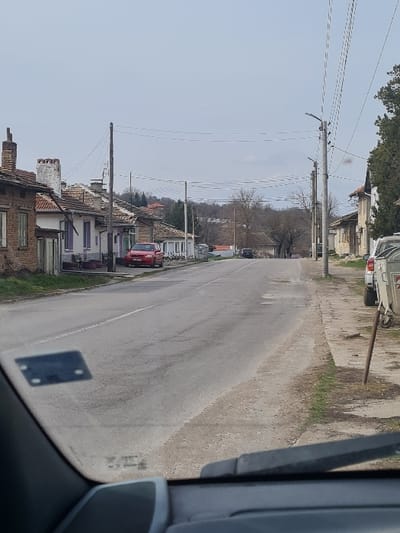 The image presents a static, forward-facing view from a vehicle, looking down a two-lane asphalt road in the city of Blagoevgrad, Bulgaria, under an overcast grey sky. The road, exhibiting surface wear, extends straight into the background, narrowing with perspective.
On the left side of the road, a series of residential buildings are visible. The closest structure features exposed red brickwork and a prominent brick chimney. Adjacent to it stands a white house with purple window trim and a tiled roof. A red sedan is parked diagonally on the dirt shoulder in front of this house, oriented towards the road. Multiple wooden utility poles carrying overhead lines line this side of the street.
On the right side of the road, a dirt and gravel shoulder with sparse vegetation runs parallel to the asphalt. Concrete utility poles equipped with streetlights and carrying numerous overhead lines are spaced along this side. The rear section of a white vehicle, possibly a minivan or SUV, is partially visible parked on the shoulder further down the road. Beyond the immediate structures, the background reveals hills populated with leafless deciduous trees, suggesting a late winter or early spring season.
The immediate foreground displays components of the vehicle from which the image was captured, including a portion of the dashboard, the windshield with a dark rectangular object (possibly a sticker or device) affixed to its lower-left area, and a windshield wiper blade positioned at the bottom edge of the frame. No individuals are present or engaged in any discernible actions within the captured scene.