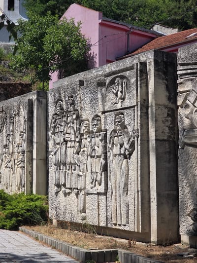 This image shows a bas-relief sculpture on a stone wall in the city of Golyamo Belovo, Bulgaria. The sculpture depicts a procession of figures, appearing to be men in uniform with stern expressions. They are rendered in a stylized, almost blocky manner, with distinct clothing and facial features. The wall itself is made of rough-hewn stone blocks and appears weathered. In the background, there are buildings, one with a pink facade and another with a red tiled roof, set against a backdrop of lush green trees and foliage. The foreground shows a paved walkway and a strip of dry grass and green bushes along the base of the wall, bordered by a concrete curb. The lighting suggests it is a sunny day, casting shadows on the sculpture and the ground.