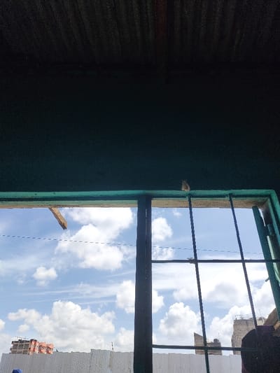 The scene depicts an exterior view through an interior window, likely situated in Juja ward, Kenya. The foreground consists of a dark, textured ceiling, possibly corrugated metal, and the top section of a window frame painted a teal or dark green color. A small, light-colored object is positioned on the top horizontal bar of the window frame. A broken piece of light-colored wood extends from the left side of the upper frame. The window features dark, vertical and horizontal security bars.

Beyond the window, the environment is a bright blue sky with numerous scattered white cumulus clouds, indicating daylight conditions. In the mid-ground, a long, white corrugated barrier or fence runs horizontally. Above this barrier, multiple multi-story buildings are visible in the distance. To the left, a building under construction shows exposed rebar and red-brown elements. To the right, another building appears partially completed or fully constructed, with darker exterior tones. A thin, dark line, possibly a utility wire, traverses the sky horizontally. The bottom edge of the image shows indistinct dark and blue fabric, suggesting the presence of an observer or objects within the interior space.