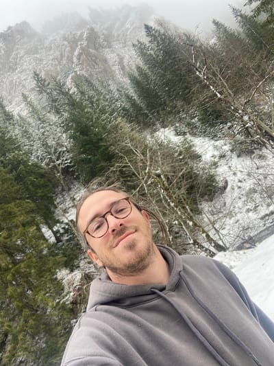 A selfie shot shows a man with glasses and a grey hoodie, seemingly standing on a snowy, forested mountain, likely in Sedlačka gora, Montenegro. The background features snow-covered trees, bare branches, and a misty mountain slope.