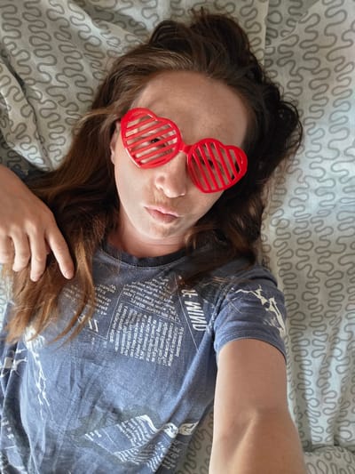 A woman with long brown hair, wearing red heart-shaped shutter glasses and a blue t-shirt with a newspaper print, lies on a bed with a grey wavy patterned duvet. Her head is turned to the side, and her left hand rests on her chest. The scene is indoors, likely in a bedroom in Буренка, Russia.