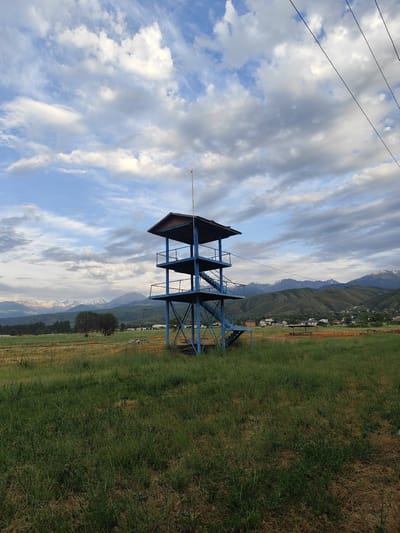 The image depicts an outdoor scene with a blue, multi-level observation tower as the central subject. The tower has a metal frame, stairs, and a roof. It is situated in a grassy field with some dry patches. In the background, there are rolling green hills with a small village nestled among them. Further in the distance, snow-capped mountains are visible under a cloudy sky with patches of blue. Power lines stretch across the upper right portion of the image. The overall impression is a rural or semi-rural landscape with a man-made structure offering a vantage point.
