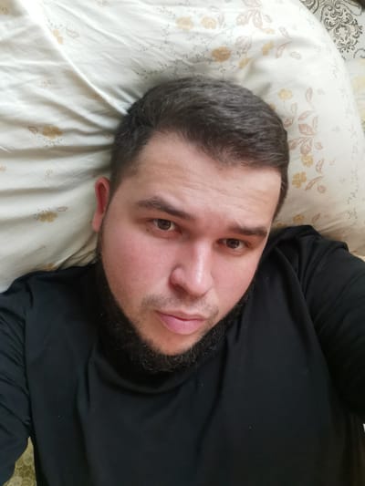 This is a selfie of a man lying down on a patterned pillow. He has short, dark hair, a beard, and is wearing a black t-shirt. The pillow has a floral pattern with yellow flowers and green leaves. The man's face is centered in the frame, and he is looking directly at the camera. His expression appears neutral. The lighting suggests it might be daytime, possibly indoors. There is no visible text in the image. The location is identified as Serov, Russia.