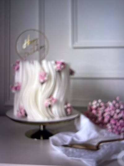 The image displays a birthday cake decorated with white frosting with wavy patterns and adorned with small pink flowers. A circular cake topper with gold lettering reads "Happy Birthday". The cake is situated on a cake stand. To the right, a bunch of pink flowers is arranged on a white cloth, partially covering an open book. The background features a white wall with molding. The scene appears to be indoors, likely in a domestic setting within the city of Beșalma, Moldova. No people are visible in the frame.