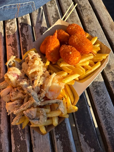 Two open, disposable paper cartons containing fried food are positioned on a weathered, slatted wooden surface outdoors in Monterosso al Mare, Italy. The carton on the left holds a substantial portion of assorted breaded and fried seafood, including calamari rings, small whole fish, and shrimp, layered over french fries. The carton on the right contains french fries along with six brightly orange, breaded, oval-shaped fried items, one of which distinctively resembles a fried crab claw. Two light-colored wooden chopsticks are visible on the wooden surface, resting diagonally above the right carton. Strong, directional sunlight illuminates the scene, casting pronounced shadows and highlighting the texture of the food and the wooden surface. A partial view of blue denim fabric is present in the upper left corner, indicating the immediate proximity of a person.