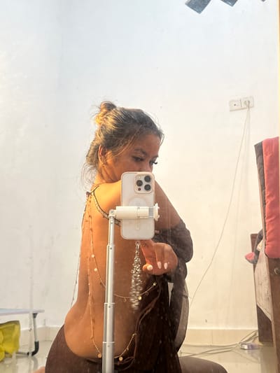 A selfie of a woman in a mirror reflection in Celuk, Indonesia. She holds a white iPhone with a tripod stand, obscuring part of her face. She wears a brown top and multiple layered necklaces. Her hair is tied up. The background includes a white wall with an electrical outlet, a shelf with pink clothing, and the corner of a white shelf.