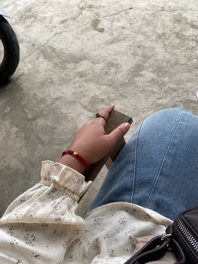 A close-up, eye-level shot shows a person's left arm and hand resting on a wooden surface. The person is wearing a light-colored, floral-patterned long-sleeved shirt with ruffled cuffs and blue denim jeans. A red beaded bracelet with a gold charm is visible on the wrist, and a gold ring with a dark stone is on the index finger. The background is a textured concrete surface, possibly a sidewalk or ground. The scene is outdoors in a non-descript urban area, likely Kutalhoksukon, Indonesia, suggested by the presence of a motorcycle wheel partially visible in the upper left corner. The lighting is natural and diffused, indicating it is daytime. There are no other people or direct interactions visible in the frame.