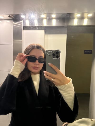 A female subject is depicted taking a selfie in a reflective indoor environment, identified as being in Akulovo (Gp Kubinka), Russia. The subject, with shoulder-length dark brown hair, wears a cream-colored ribbed turtleneck sweater under an open dark coat. She holds a smartphone with a gray, irregularly shaped case in her left hand, angled towards herself. Her right hand adjusts a pair of dark, rectangular-framed sunglasses, which are positioned partially down her nose. A thin band ring is visible on the ring finger of her left hand. The background consists of highly reflective wall panels, consistent with an elevator interior or mirrored hallway. Overhead, multiple recessed light fixtures illuminate the space. Reflections of a dark blue sign displaying "MATE P" and a light-colored object with "OLEX" text are visible on the reflective surfaces.