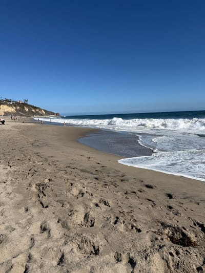 The image depicts a wide, sandy beach in Malibu, California, United States of America, on a clear, sunny day. The foreground is dominated by the textured sand, showing numerous footprints and some scattered seaweed. The ocean, with its characteristic blue hue, occupies the middle ground, featuring moderately sized waves breaking on the shore, creating white foam. In the background, a rugged cliff face rises, topped with houses and palm trees, indicative of a coastal residential area. Several people are scattered across the beach, some walking, others sitting or playing near the water's edge. The sky is a deep, cloudless blue.