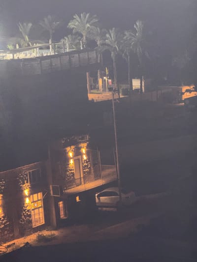 The image depicts an outdoor scene at night in Tiberias, Israel. In the foreground, a two-story building constructed of rough-hewn stone is visible, illuminated by several warm-toned lights on its exterior walls and through its windows. One window on the lower level features a stained-glass design. An air conditioning unit is mounted on the exterior wall. A balcony with a decorative railing extends from the upper level of the building. A white car is parked in front of the building. In the middle ground, there is a power pole with wires. Further in the background, palm trees are scattered across the landscape, and there are structures that appear to be part of a resort or hotel, including a shaded seating area with lounge chairs and umbrellas. The overall atmosphere is dark and atmospheric, with artificial lighting creating highlights and shadows.