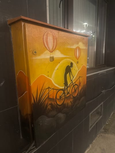 The image shows a painted electrical box on a dark tiled wall in Belogradchik, Bulgaria. The painting depicts a silhouette of a person riding a bicycle against a vibrant sunset with two hot air balloons in the sky. The artwork is done in warm colors of orange, yellow, and red, with dark silhouettes for the landscape and figures. The electrical box is mounted on a wall that has a window or opening to its right, reflecting some distant lights. The foreground of the painting features a grassy landscape with the silhouette of a fence.