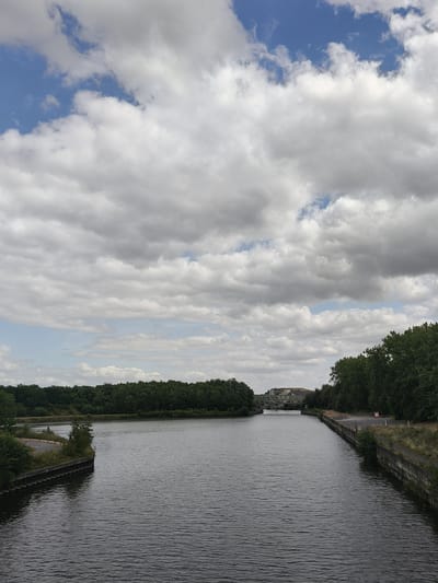 The image depicts a wide canal, with the water surface showing ripples. On the left bank of the canal, a paved path curves away, bordered by a low concrete wall and vegetation. The right bank is also lined with a concrete wall and has a grassy embankment above it, with a road running alongside. Dense green trees form a backdrop along both banks, extending into the distance. In the far distance, a bridge crosses the canal, with buildings visible on the horizon beyond the bridge. The sky is filled with a mix of white and gray clouds, with patches of blue sky showing through. The scene is captured from an elevated vantage point, looking down the length of the canal. This is likely the Canal de Courcelles in Dourges, France, given the presence of canals and bridges in the area. No people are visible in the image.
