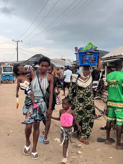 In Oko Amakom, Nigeria, a group of people are walking on a dirt road under an overcast sky. In the foreground, a young woman in a striped dress carries a child in her arms. The child is reaching towards the woman's face. The woman is wearing a patterned wrap around her waist and white flip-flops. To her right, a younger girl in a pink top and shorts walks ahead. Behind the woman carrying the child, another woman wears a headscarf and a patterned dress, with a blue basket full of items balanced on her head. To the far right, a person in a bright green shirt and shorts walks away from the camera. In the background, there are buildings with corrugated metal roofs, a blue tricycle, and other people milling about. Power lines are visible overhead, supported by a utility pole. The ground is dusty and uneven, with some debris scattered about.