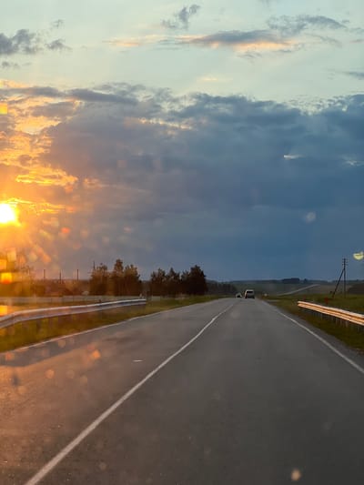 The image depicts a road at dusk, with the sun setting on the left side, casting a warm glow on the clouds and creating lens flare effects. The road, which appears to be a highway, has two lanes and a white line marking the center. A guardrail runs along the right side of the road. Several cars are visible on the road, driving in the same direction as the viewer. In the background, there are trees, some buildings, and utility poles. The sky is a mix of light blue and dark clouds, suggesting changing weather conditions. The scene is likely captured from a moving vehicle. The location is described as the Чырвонаслабодскі сельскі Савет in Belarus.