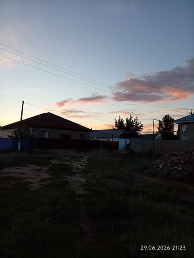 This is a landscape photograph taken at dusk in Qobda, Kazakhstan. The scene depicts a rural residential area with several houses visible. In the foreground, a dirt path winds through tall, dark green grass.
The main subject is a long, single-story house with a dark-colored, tiled roof and lighter-colored walls. It is bordered by a dark fence. To the left of this house, a tall wooden pole stands. Further into the background, other houses and fences are visible, including one with a blue roof and satellite dishes mounted on its exterior. Several trees are scattered throughout the scene.
The sky is a prominent feature, showing the transition from day to night. The upper portion is a clear, pale blue, while lower down, scattered clouds are tinged with pink and orange from the setting sun. Three thin power lines are strung across the sky, adding a man-made element to the natural landscape. The overall mood is serene, with the soft light of dusk casting shadows and creating a peaceful atmosphere. The image captures a quiet evening in a rural settlement.
The bottom right corner of the image contains visible text: "29.06.2026 21:23".