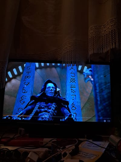 The image shows a television screen displaying a scene from a movie. On the screen, a skeletal figure in a dark hood and armor is visible. The figure is positioned between two tall, blue, stone-like structures adorned with glowing, alien-like symbols. The background of the screen shows a dimly lit, circular, chamber-like setting with arches. Below the television screen, various electrical cables and power strips are visible, suggesting a domestic setting. A portion of a warning label with Polish text is also visible in the foreground, indicating the location of this scene is likely within a private residence in Mława, Poland.