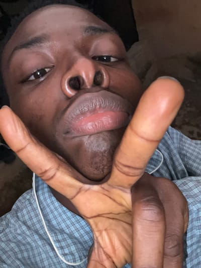 A close-up, indoor photograph shows a dark-skinned individual with their face at an angle. Their right hand is raised, obscuring part of their face with the index and middle fingers extended in a V-shape. The person is wearing a blue and white checkered shirt. No specific location in the city of Kirana, Nigeria, is identifiable.