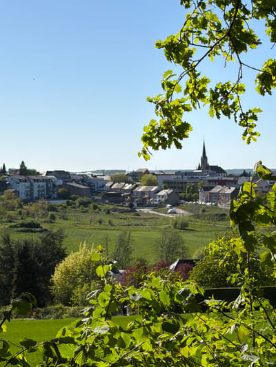 The image depicts a scene in Pétange, Luxembourg, viewed from an elevated perspective through tree foliage. A town stretches along a line, featuring buildings and a church spire. Green spaces, possibly fields or parkland, adjoin the built environment. The sky is clear blue. The image appears to be rotated counterclockwise.