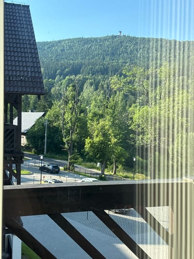 This media file captures a scenic view of a wooded mountainside in Szklarska Poręba, Poland. The foreground features a dark-tiled roof and a wooden railing, suggesting a vantage point from a building, possibly a balcony or window. Sunlight streams through horizontal blinds, creating a striped pattern across the image.
Dominating the background is a dense, green forest covering a gently sloping mountain. A clear blue sky is visible above the treeline. A narrow road winds through the trees, with a few cars visible, including a dark-colored sedan and a white vehicle. Yellow barriers line sections of the road, and a streetlamp stands by its side. The presence of these elements indicates a rural or semi-rural location, likely a tourist area given the road and proximity to nature.
The lighting suggests it is daytime, possibly late morning or early afternoon, given the bright sunlight. The weather appears to be clear and pleasant, conducive to outdoor activities. The scene evokes a sense of tranquility and natural beauty. No people are clearly visible, and no text is discernible in the image. The overall impression is one of a peaceful, sunlit day in a mountainous landscape.
